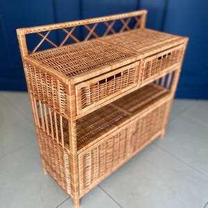 May include: A light brown wicker storage cabinet with two drawers, a shelf, and a cabinet door. The cabinet has a decorative crisscross design on the top and is made of woven wicker.