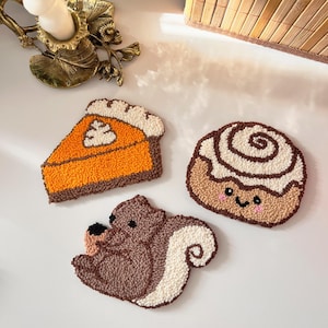 May include: Three handmade, textured coasters: a slice of pumpkin pie, a cinnamon roll with a smiling face, and a squirrel. The pie is orange with a brown crust, the roll is tan with white icing, and the squirrel is brown and white.