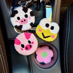 May include: A collection of four colorful, handmade car coasters. Designs include a cow, a pig, a frog, and a flower. The coasters are made of a textured, looped material and are placed in a car's center console.