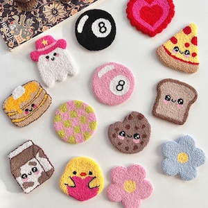 Op de afbeelding: Een collectie getufte onderzetters met diverse vormen, waaronder een zwarte achtbal, een roze bal, een hart, een pizzapunt, een spookje met een roze hoed, pannenkoeken, een geblokte cirkel, een koekje, toast, een melkpak, een kuikentje en bloemen.