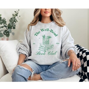 Puede incluir: Una sudadera gris con el texto "The Well-Read Book Club" en letras verdes. El diseño incluye un vaso de cóctel con una rodaja de lima y una pila de libros. El texto "Est. 2020" también está incluido en el diseño.