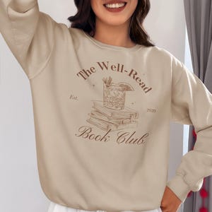 Puede incluir: Una sudadera beige con un diseño gráfico marrón que dice "The Well-Read Book Club Est. 2020" con un dibujo de una copa de cóctel y libros.
