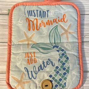 May include: A quilted pot holder with a light blue background, coral trim, and a coral loop. The design features a mermaid tail, starfish, and the text "INSTANT Mermaid JUST ADD Water". A wooden button is attached.