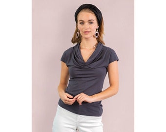 Puede incluir: Una blusa gris oscuro con cuello de cisne y mangas cortas. La blusa se combina con pantalones blancos. La persona lleva una diadema negra y pendientes. El fondo es de color rosa claro liso.