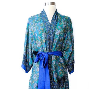 May include: A teal floral print robe with a blue waist tie. The robe features a detailed floral pattern in shades of teal, blue, and white. The robe is displayed on a mannequin against a white background. The robe has long sleeves and a knee-length hem.
