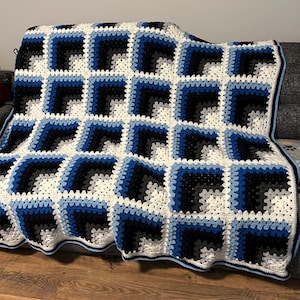 May include: A handmade crochet blanket in a geometric pattern. The blanket features squares in white, black, gray, and shades of blue. The blanket is draped over a couch, showcasing its texture and design. The edges are finished with a blue border.