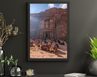 Puede incluir: Impresión enmarcada del Tesoro en Petra, Jordania. La antigua estructura está tallada en un acantilado de arenisca. Los camellos descansan en primer plano, con turistas y un cielo brillante visibles.