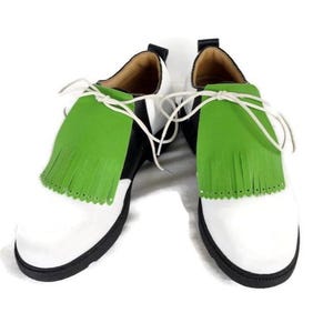 Lime green apple golf shoe kilties on a pair of mens black and white saddle style golf shoes with white shoe laces and black soles. Kilties have 15 fringes each with a single perforation in the point. Front view. White background.