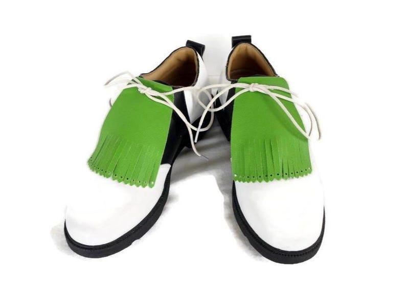 Lime green apple golf shoe kilties on a pair of mens black and white saddle style golf shoes with white shoe laces and black soles. Kilties have 15 fringes each with a single perforation in the point. Front view. White background.