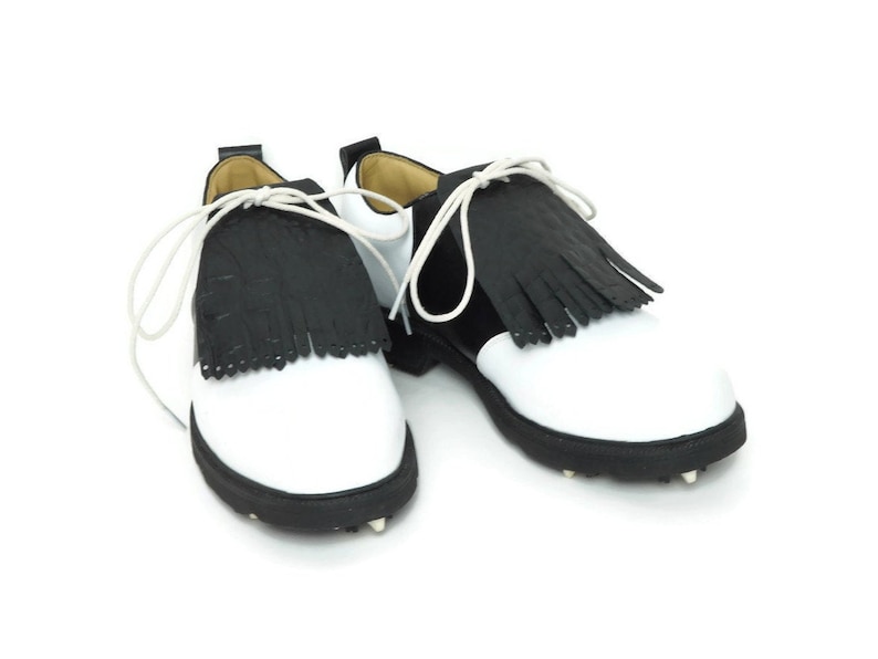 Black alligator embossed leather shoe fringes on a pair of black and white saddle shoes for men, tied with white shoe laces, black rubber soles. Classic kilties have 16 fringes with a single perforation near each tip. Front view.