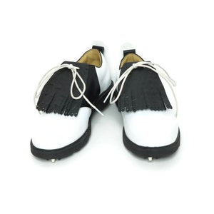 Black alligator embossed leather shoe fringes on a pair of black and white saddle shoes for men, tied with white shoe laces, black rubber soles. Classic kilties have 16 fringes with a single perforation near each tip. Front view, shoes apart.