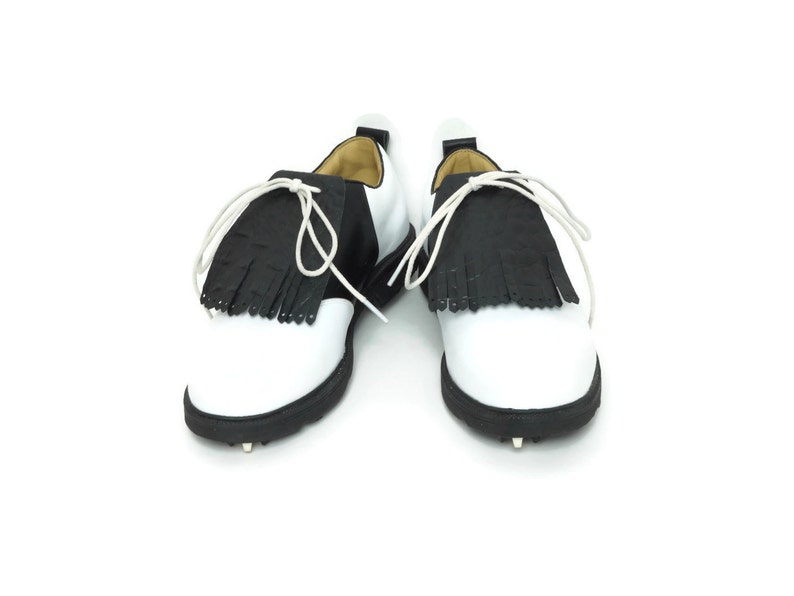 Black alligator embossed leather shoe fringes on a pair of black and white saddle shoes for men, tied with white shoe laces, black rubber soles. Classic kilties have 16 fringes with a single perforation near each tip. Front view, shoes apart.