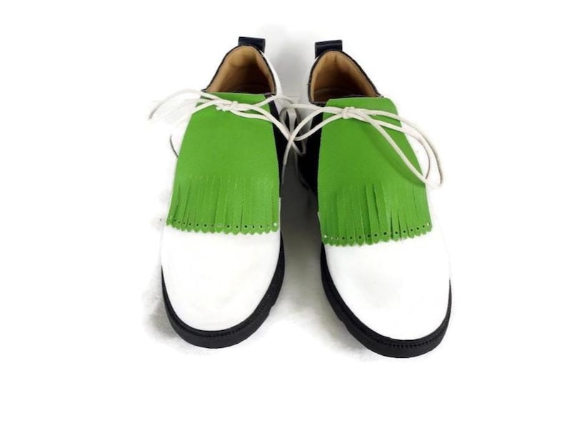 May include: A pair of white and black shoes with a green fringe detail. The shoes have white laces and a black sole. The toe of the shoe is white, and the upper part is black.
