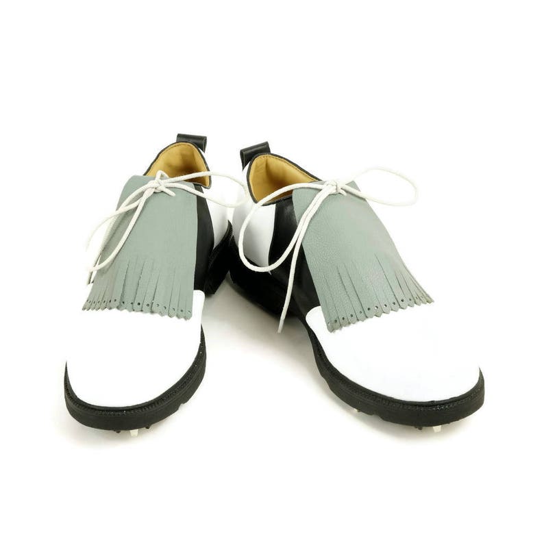 May include: A pair of white and black golf shoes with grey fringe accents. The shoes have a white leather upper with a black leather trim around the toe and heel. The shoes have a grey fringe that hangs down from the top of the shoe. The shoes have a white rubber sole with black cleats.