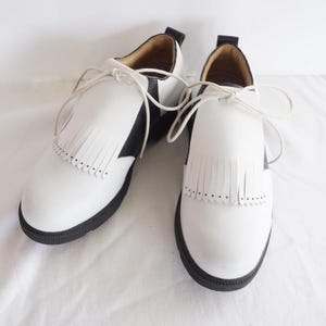 May include: A pair of white leather golf shoes with black trim and fringe detailing. The shoes have laces and a black rubber sole.