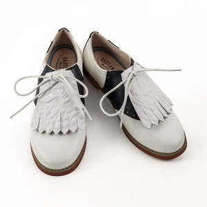 May include: White leather saddle shoes with black trim and fringe detail. The shoes have white laces and the brand name "Eastland" is printed on the side.