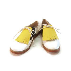 May include: Pair of white leather shoes with yellow fringe detail and brown soles. The shoes have black and white accents and white laces. A stylish and unique footwear option.