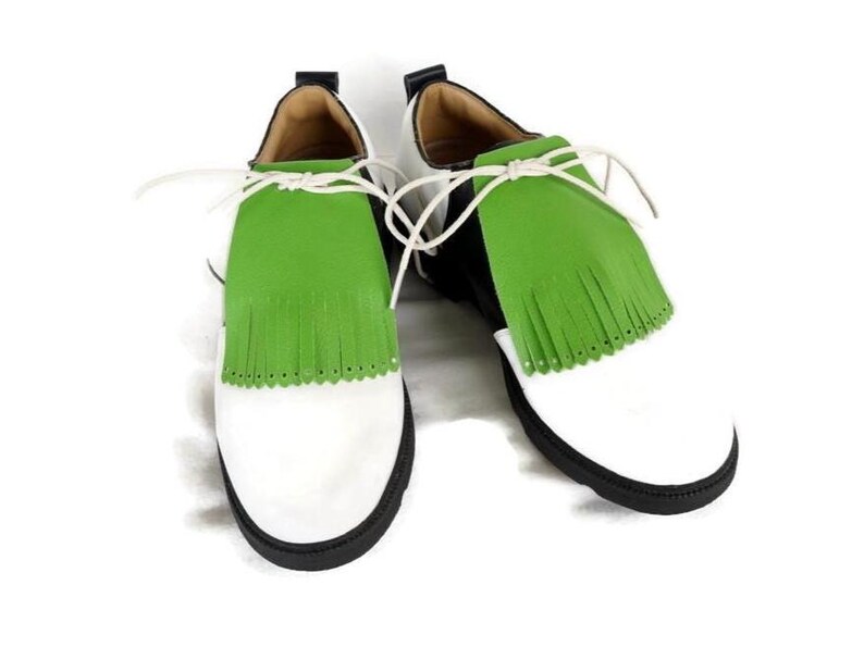 Lime green apple golf shoe kilties on a pair of mens black and white saddle style golf shoes with white shoe laces and black soles. Kilties have 15 fringes each with a single perforation in the point. Front view. Solid white background.