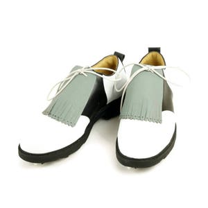 May include: A pair of white and black golf shoes with grey fringe accents. The shoes have a black rubber sole with small spikes.