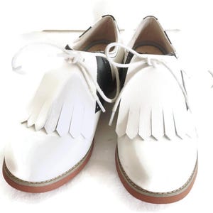 May include: Pair of white leather shoes with fringe detailing and black accents. The shoes have a classic lace-up design and a brown sole. A stylish and unique footwear option.