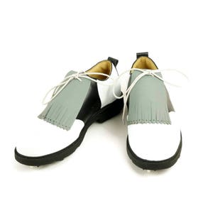May include: A pair of white and black golf shoes with grey fringe accents. The shoes have a white toe cap and a black heel.