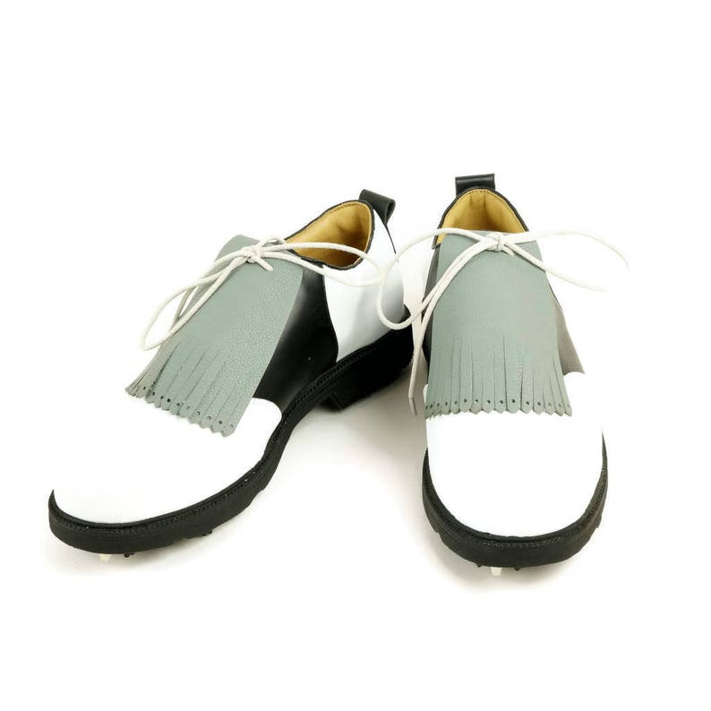 May include: A pair of white and black golf shoes with grey fringe accents. The shoes have a white toe cap and a black heel.