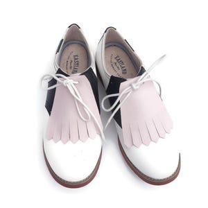 May include: A pair of two-tone saddle shoes with white and black leather, featuring a pink fringe detail and white laces. The shoes have a red sole and the brand name "Eastland" is visible on the insole.