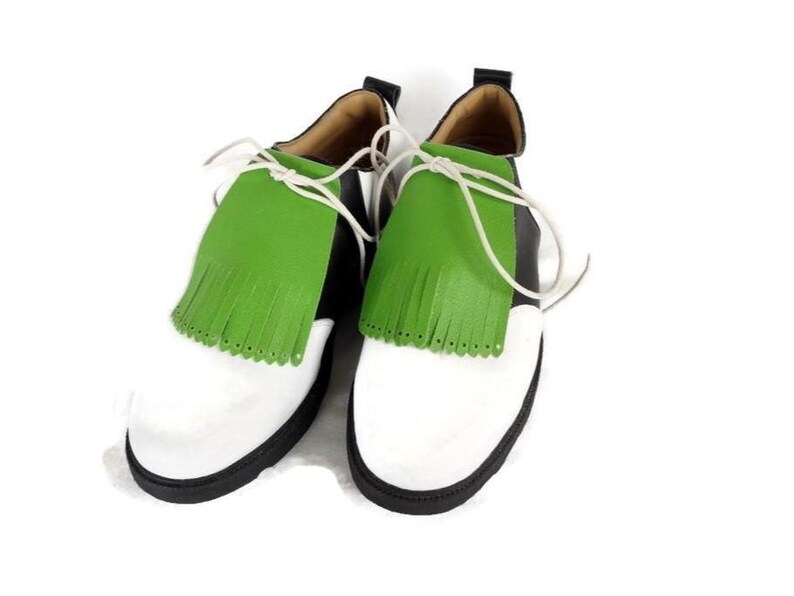 Lime green apple golf shoe kilties on a pair of mens black and white saddle style golf shoes with white shoe laces and black soles. Kilties have 15 fringes each with a single perforation in the point. Front view. White background.