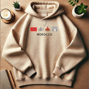 May include: A beige hoodie featuring a graphic of a Moroccan flag, teapot, tagine, and archway. The text "MOROCCO" is printed below the graphic.