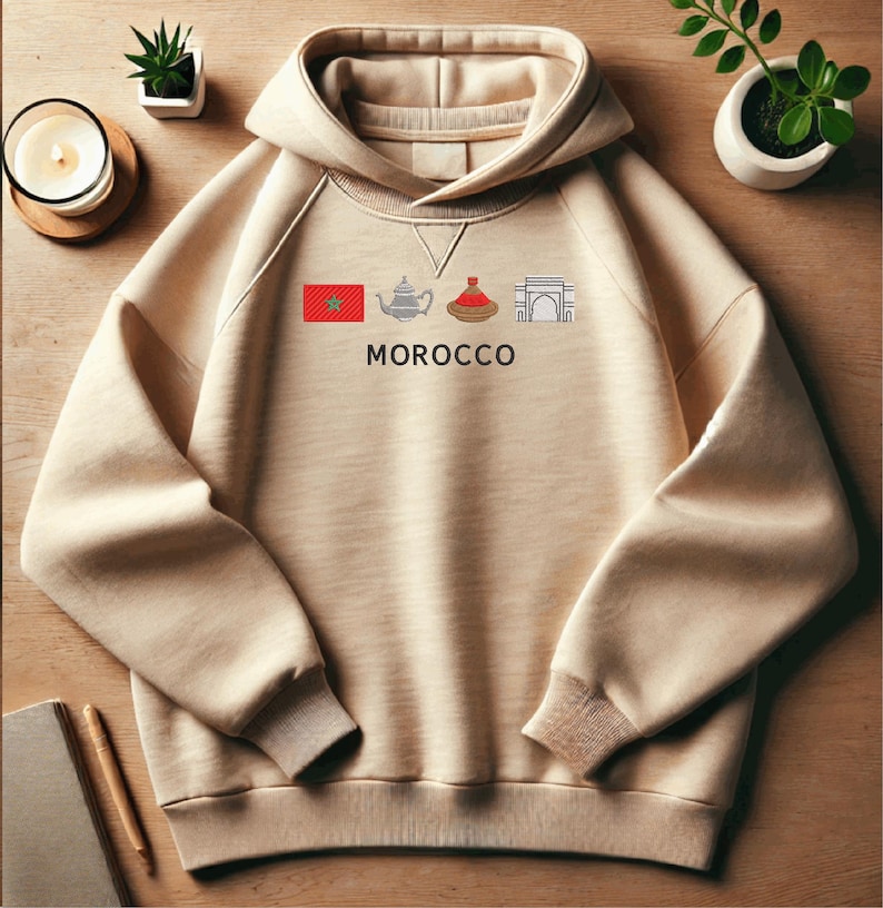 May include: A beige hoodie featuring a graphic of a Moroccan flag, teapot, tagine, and archway. The text "MOROCCO" is printed below the graphic.
