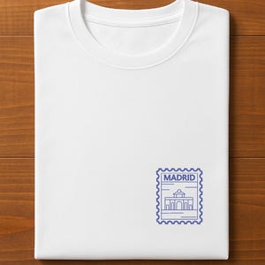 May include: A folded white t-shirt with a blue postage stamp design featuring the word "MADRID" and a building illustration. The shirt is displayed on a wooden surface.