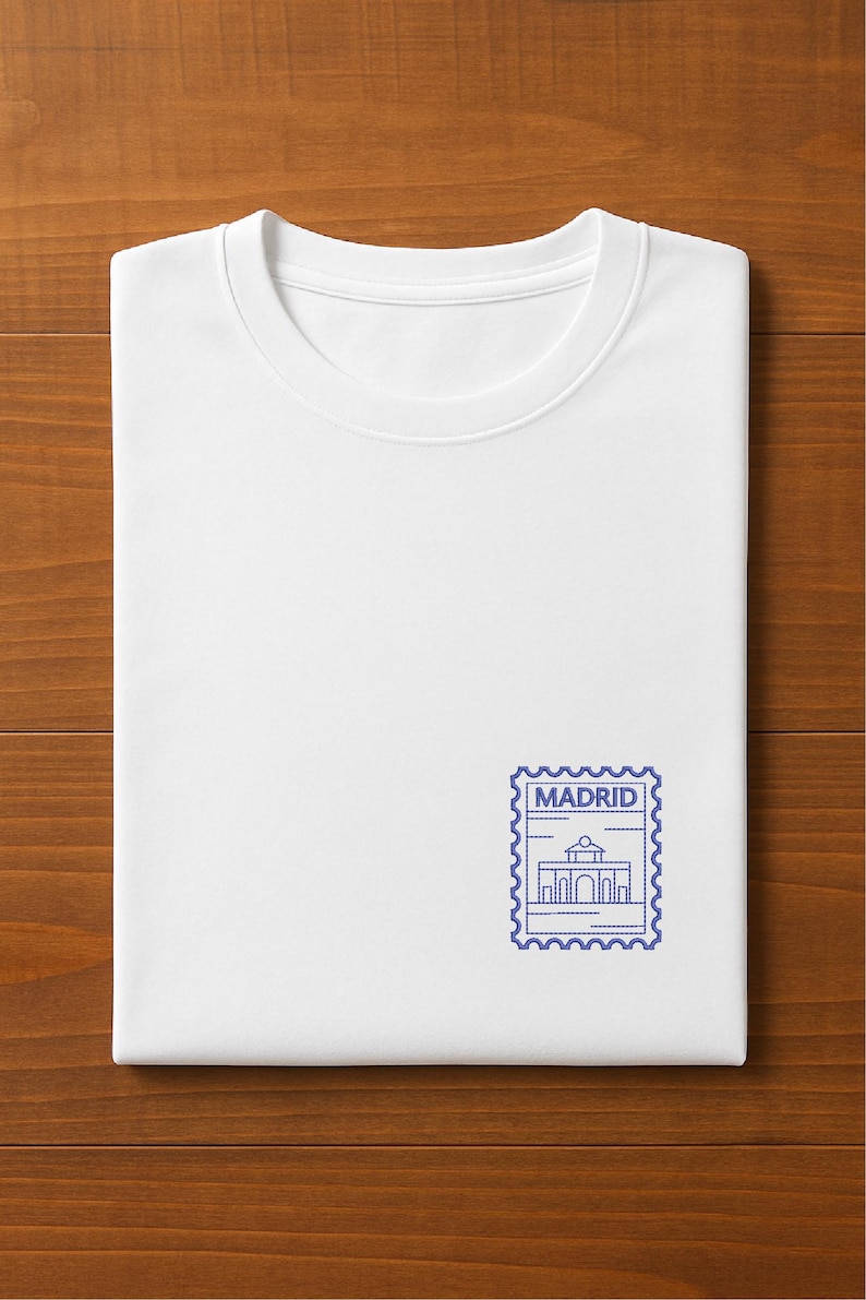 May include: A folded white t-shirt with a blue postage stamp design featuring the word "MADRID" and a building illustration. The shirt is displayed on a wooden surface.