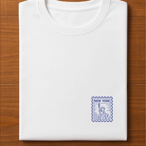 K&ouml;nnte beinhalten: Gefaltetes wei&szlig;es T-Shirt mit blauem New York-Stempeldesign. Der Stempel zeigt die Freiheitsstatue und den Text "NEW YORK". Das T-Shirt liegt auf einer Holzoberfl&auml;che.