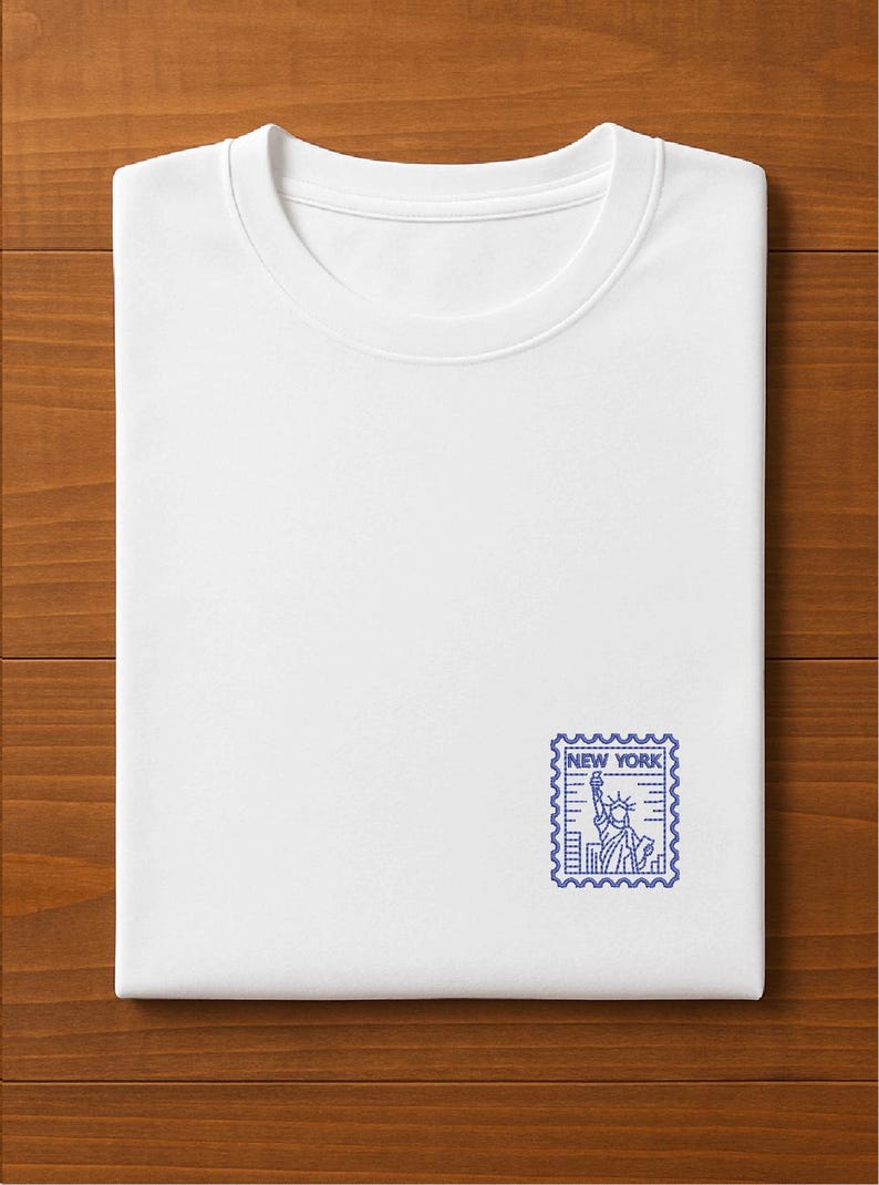 K&ouml;nnte beinhalten: Gefaltetes wei&szlig;es T-Shirt mit blauem New York-Stempeldesign. Der Stempel zeigt die Freiheitsstatue und den Text "NEW YORK". Das T-Shirt liegt auf einer Holzoberfl&auml;che.