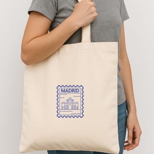 May include: Cream-colored tote bag with a blue Madrid stamp design. The stamp features a building illustration and the city name. The bag is held by a person wearing a gray t-shirt and blue jeans.