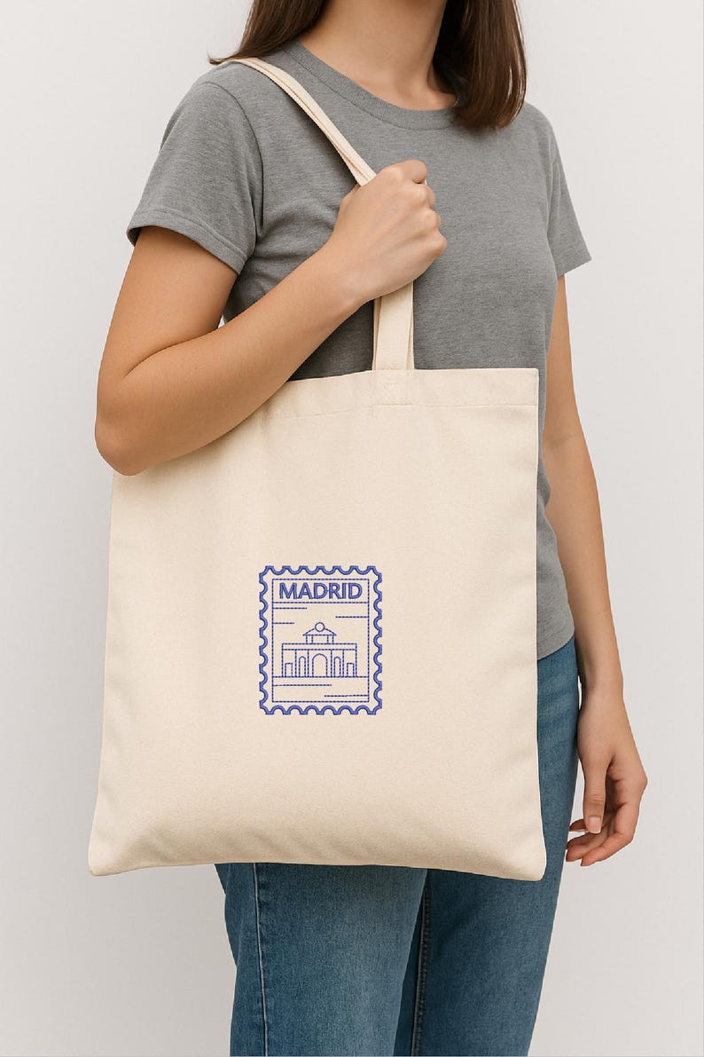 May include: Cream-colored tote bag with a blue Madrid stamp design. The stamp features a building illustration and the city name. The bag is held by a person wearing a gray t-shirt and blue jeans.