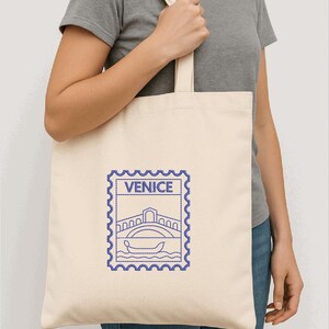 以下が含まれることがあります： クリーム色のトートバッグ。青色の刺繍で橋とゴンドラのデザイン、上に「VENICE」の文字。グレーのTシャツとブルージーンズを着た人がバッグを持っています。