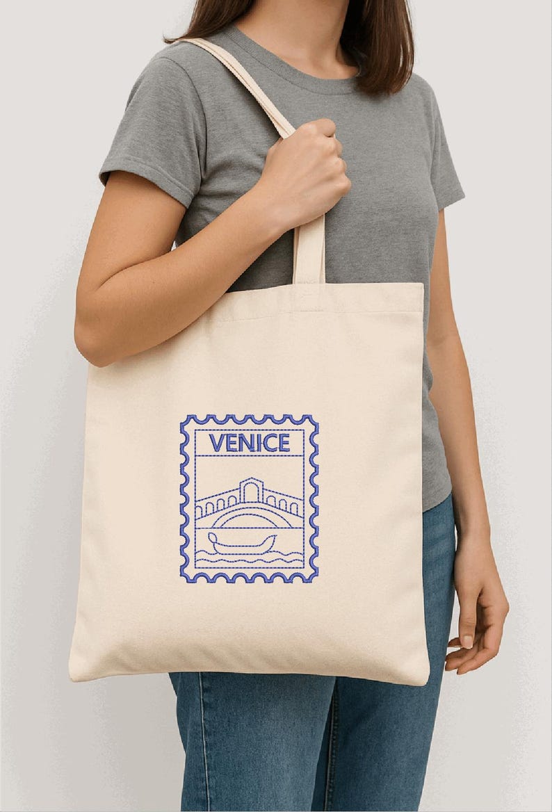 以下が含まれることがあります： クリーム色のトートバッグ。青色の刺繍で橋とゴンドラのデザイン、上に「VENICE」の文字。グレーのTシャツとブルージーンズを着た人がバッグを持っています。