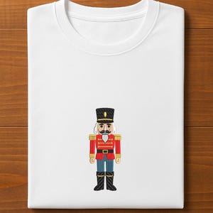 May include: White crew neck t-shirt featuring an embroidered image of a toy soldier nutcracker. The nutcracker is dressed in a red jacket, black boots, and a tall black hat. The design is centered on the front of the folded shirt.