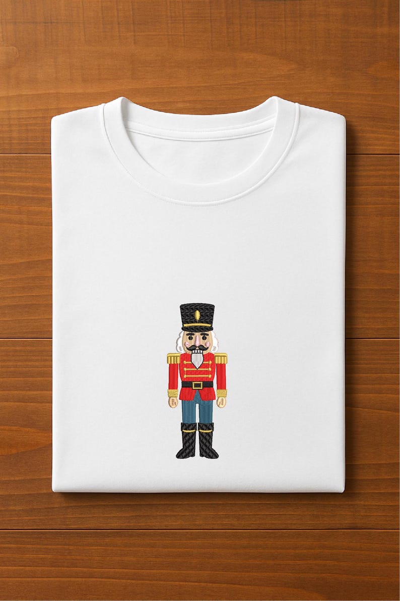May include: White crew neck t-shirt featuring an embroidered image of a toy soldier nutcracker. The nutcracker is dressed in a red jacket, black boots, and a tall black hat. The design is centered on the front of the folded shirt.
