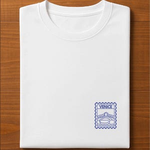以下が含まれることがあります： 青い刺繍の切手デザインが施された、折りたたまれた白いTシャツ。切手には橋とゴンドラが描かれており、「VENICE」の文字が入っています。Tシャツは柔らかい素材でできており、木製の表面に折りたたまれています。