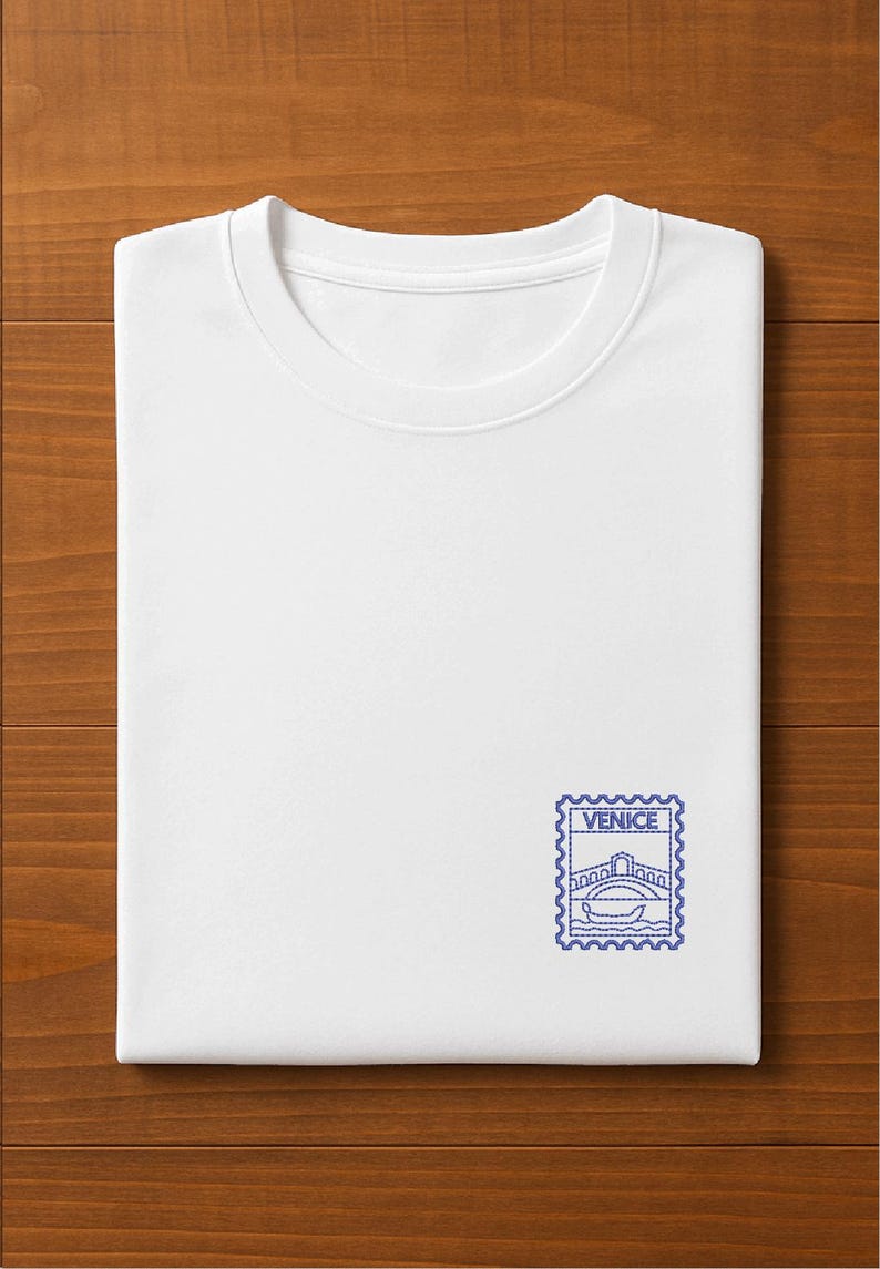 以下が含まれることがあります： 青い刺繍の切手デザインが施された、折りたたまれた白いTシャツ。切手には橋とゴンドラが描かれており、「VENICE」の文字が入っています。Tシャツは柔らかい素材でできており、木製の表面に折りたたまれています。