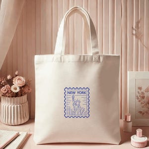 K&ouml;nnte beinhalten: Cremefarbene Canvas-Tragetasche mit blauem Briefmarken-Design, das die Freiheitsstatue und den Text "NEW YORK" zeigt. Die Tasche hat zwei Griffe und eignet sich zum Tragen von Gegenst&auml;nden.