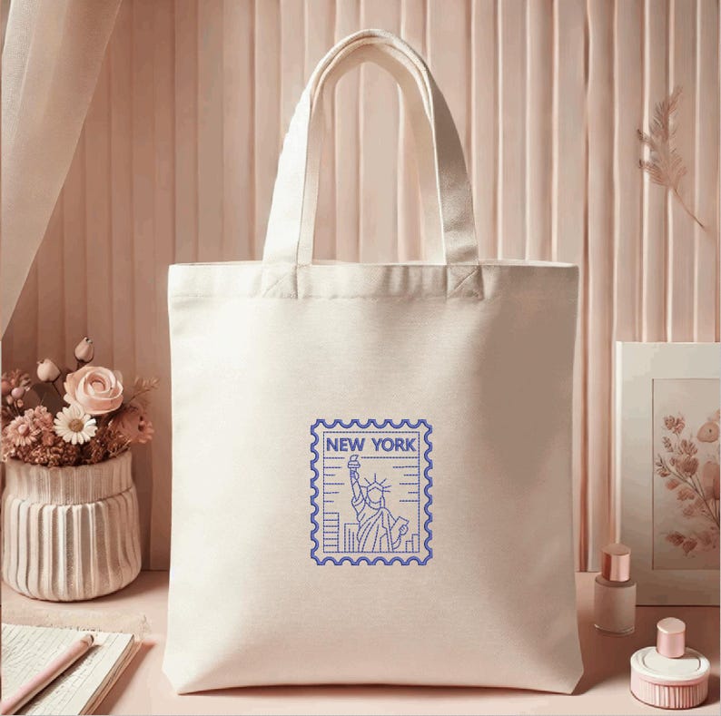 K&ouml;nnte beinhalten: Cremefarbene Canvas-Tragetasche mit blauem Briefmarken-Design, das die Freiheitsstatue und den Text "NEW YORK" zeigt. Die Tasche hat zwei Griffe und eignet sich zum Tragen von Gegenst&auml;nden.