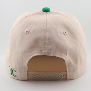 Op de afbeelding: Een beige baseballpet met een groene klep en een groene geborduurde "NC" aan de zijkant. De pet heeft een verstelbare band met een plastic gesp.