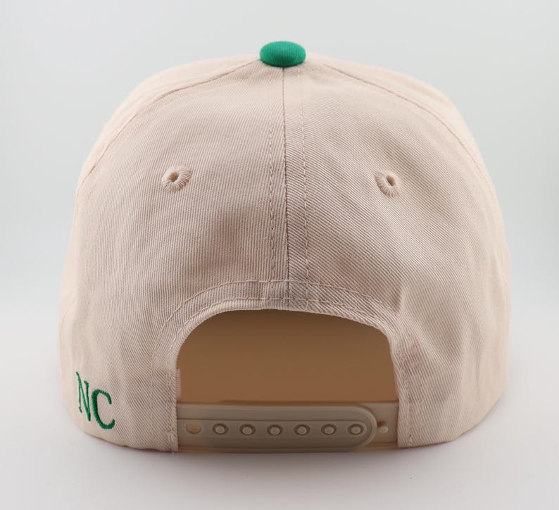 Op de afbeelding: Een beige baseballpet met een groene klep en een groene geborduurde "NC" aan de zijkant. De pet heeft een verstelbare band met een plastic gesp.
