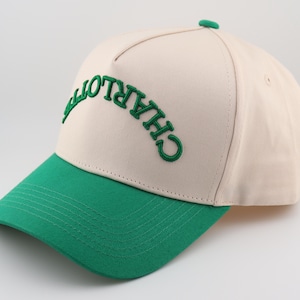 Op de afbeelding: Een beige en groene baseballpet met het woord "CHARLOTTE" in het groen geborduurd op de voorkant.