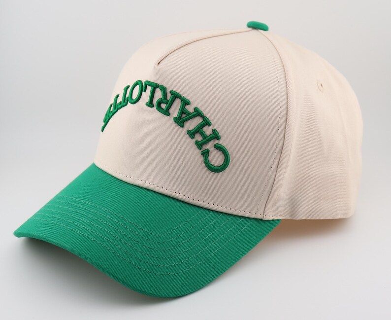Op de afbeelding: Een beige en groene baseballpet met het woord "CHARLOTTE" in het groen geborduurd op de voorkant.