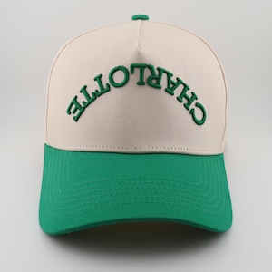 Op de afbeelding: Een beige en groene baseballpet met het woord "CHARLOTTE" in het groen geborduurd op de voorkant.
