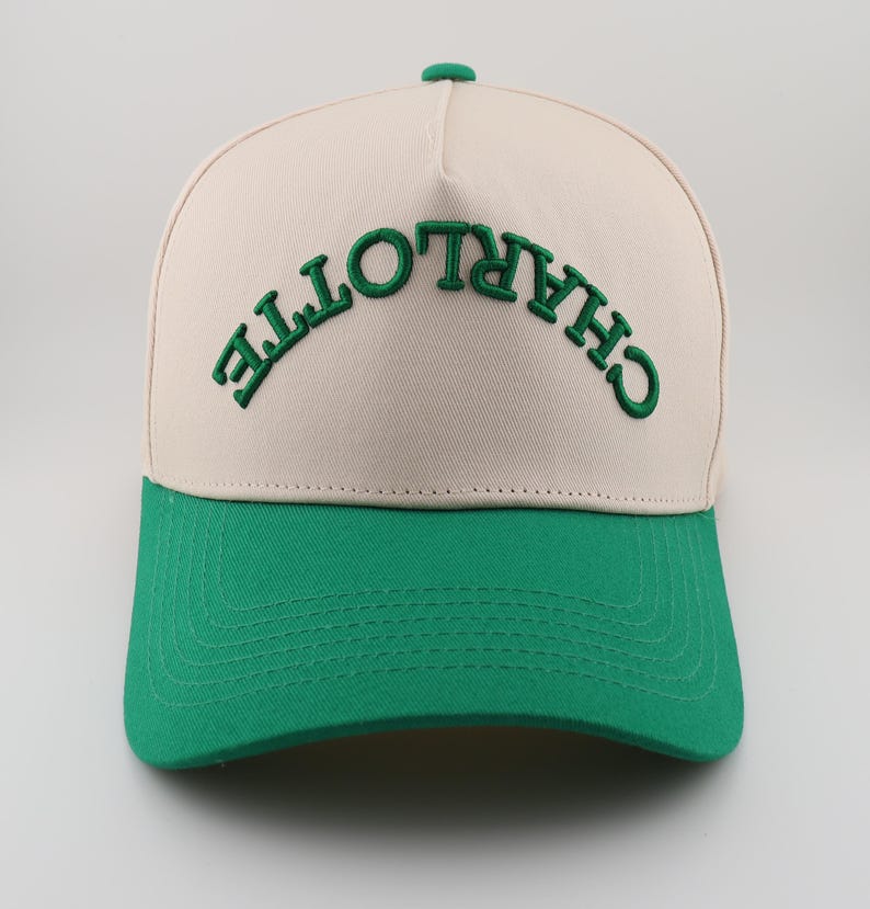 Op de afbeelding: Een beige en groene baseballpet met het woord "CHARLOTTE" in het groen geborduurd op de voorkant.
